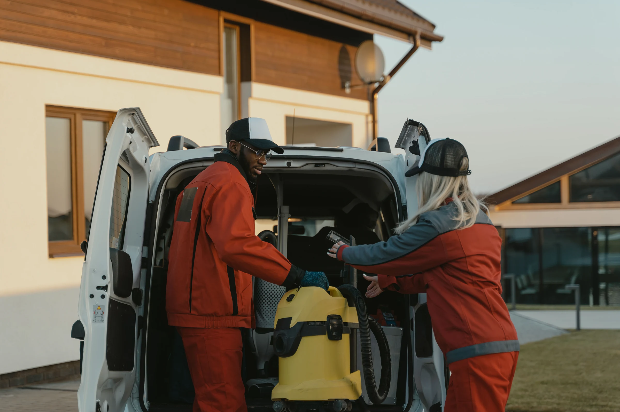 Two cleaners unloading a vacuum from a van outside a house