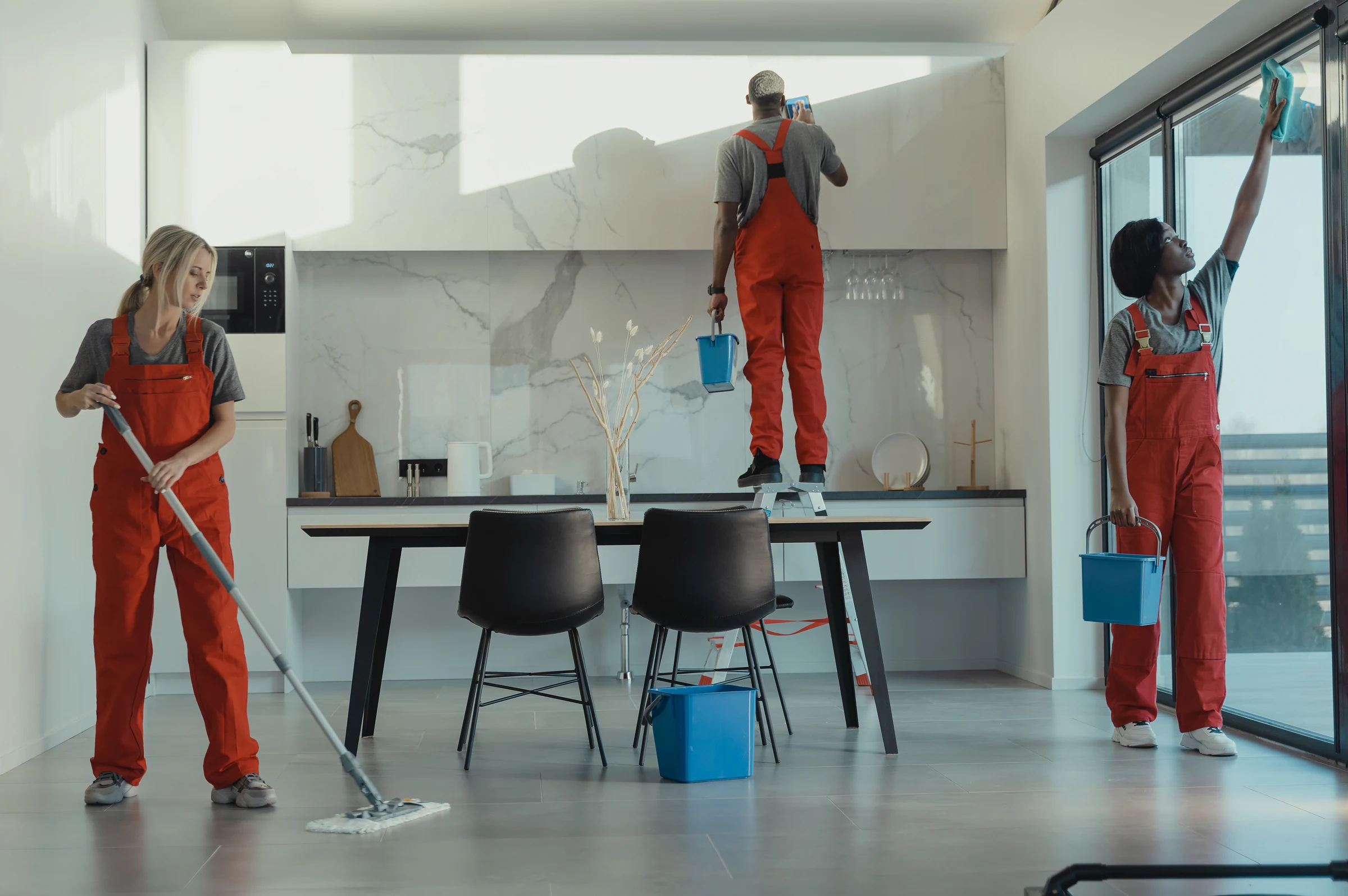 Three cleaners in overalls mopping, wiping a high wall and cleaning a window in a kitchen